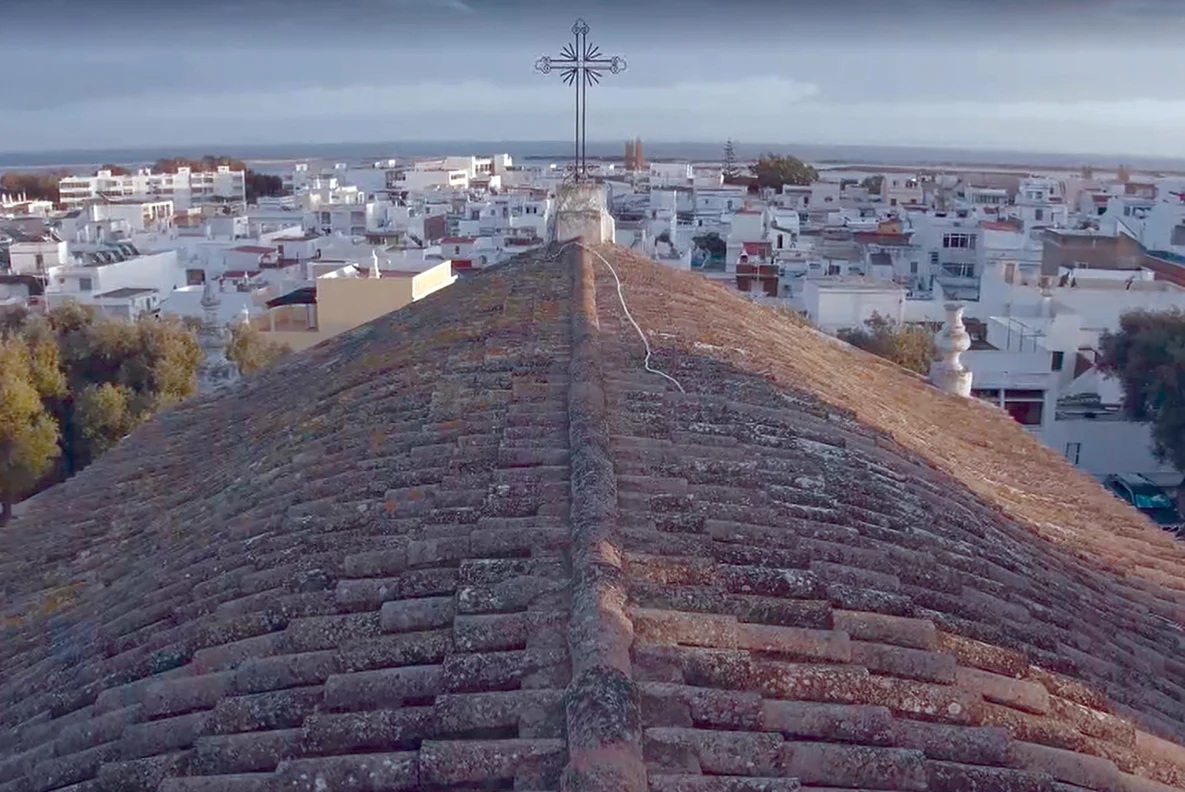 Telhado de telha de uma igreja, com cruz de ferro no cume, sobre o casario branco da Fuseta e a ria no horizonte.