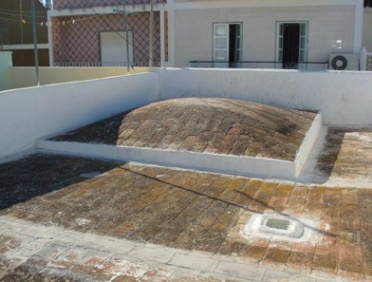 Açoteia pavimentada a tijolo com a curva de uma abóbada a emergir do terraço, entre muretes brancos e uma casa ao fundo.