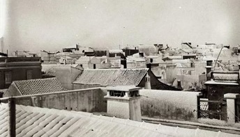 Fotografia antiga a preto e branco de telhados de telha e de uma chaminé com aberturas no remate, com o casario ao fundo.