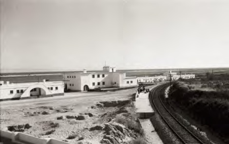 Fotografia antiga a preto e branco do Bairro dos Pescadores, com edifícios brancos junto à linha do comboio e a ria ao fundo.