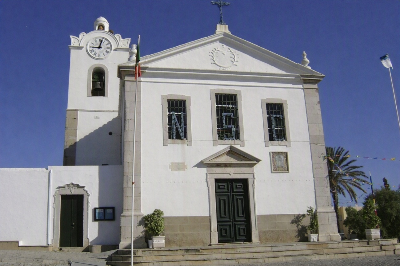 Fachada branca da Igreja Matriz da Fuseta sob céu azul, com torre sineira e relógio, frontão com cruz e as letras N, S e C nas grades das janelas