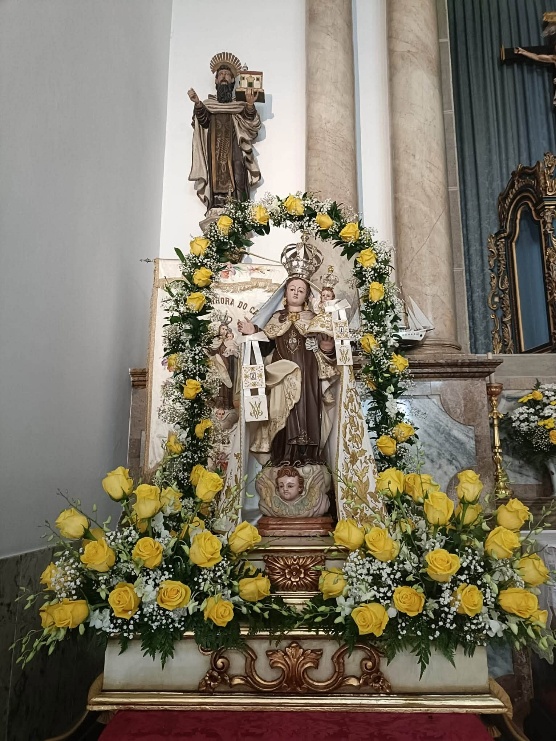 Escultura de Nossa Senhora do Carmo com o Menino, ambos coroados, num andor branco e dourado, sob um arco de rosas amarelas