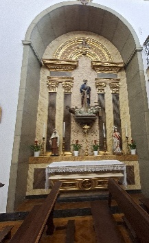 Altar lateral em arco com colunas escuras e capitéis dourados, imagem sobre mísula ao centro, duas imagens menores e vasos de flores vermelhas