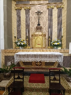 Altar com toalha de renda branca, sacrário dourado encimado por crucifixo, jarras de flores amarelas e brancas e dois genuflexórios de madeira escura