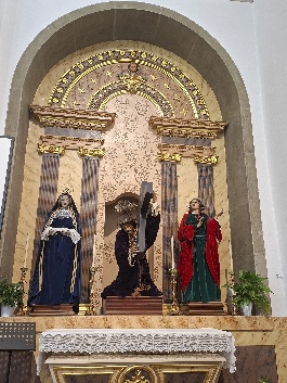 Altar lateral com imagem de Cristo de cruz às costas ao centro, entre uma figura de manto azul-escuro e outra de vermelho e verde
