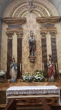Altar lateral com imagem de manto azul e o Menino ao colo sobre mísula, ladeada por outras duas imagens e jarras de flores