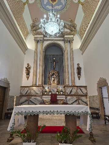 Capela-mor com altar de toalha branca, colunas de mármore, nicho dourado da Padroeira, crucifixo, lustre de cristal e teto pintado