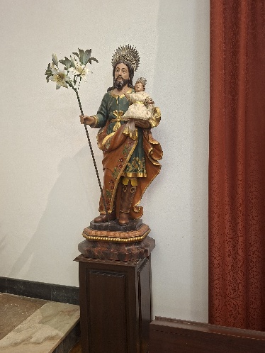 Escultura policromada de São José com o Menino ao colo e ramo de lírios, sobre pedestal de madeira junto a uma cortina vermelha
