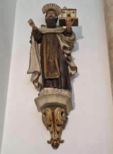 Escultura policromada de um santo barbudo, de hábito escuro e capa branca, com uma maqueta de igreja na mão, sobre mísula dourada na parede