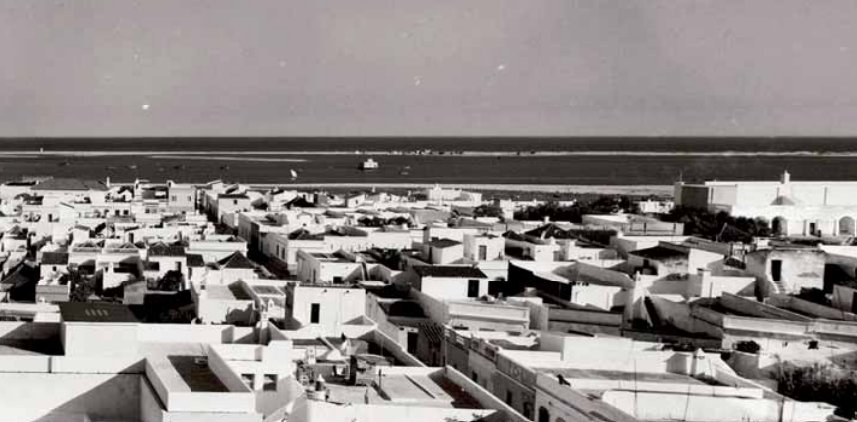 Fotografia antiga a preto e branco do casario branco da Fuseta visto do alto, com a ria, a ilha-barreira e o mar ao fundo