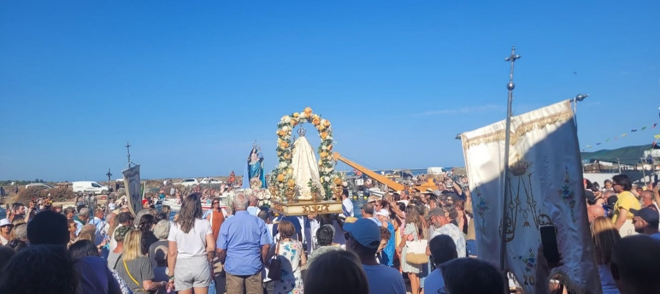 Multidão em procissão junto à ria, com o andor florido de Nossa Senhora do Carmo, visto de costas, outra imagem de manto azul e um estandarte bordado