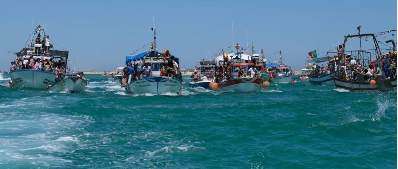 Barcos de pesca engalanados com bandeirinhas e cheios de gente navegam em águas agitadas azul-esverdeadas durante a procissão marítima