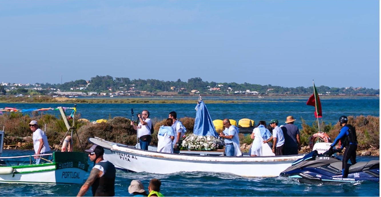 Barco branco na ria leva uma imagem de Nossa Senhora de manto azul entre flores brancas, com uma mota de água da Polícia Marítima ao lado