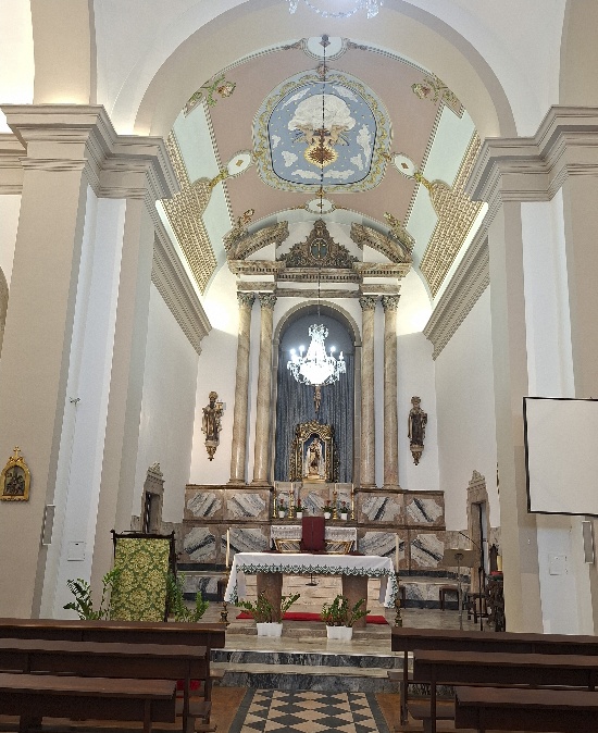 Nave da igreja vista dos bancos de madeira em direção à capela-mor, com altar de toalha branca, colunas de mármore, lustre e teto pintado