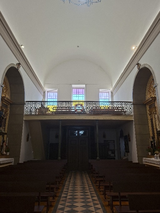 Nave vista em direção à entrada, com uma boia salva-vidas no coro-alto de balaustrada de ferro, três janelas de vidros coloridos e bancos de madeira