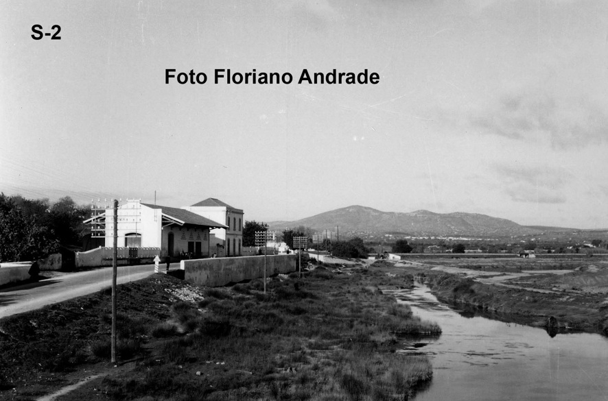 Fotografia antiga a preto e branco: estrada junto à estação de caminho de ferro, com um curso de água entre ervas e a serra ao fundo. Crédito «Foto Floriano Andrade».
