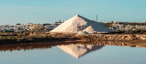 Monte de sal branco numa salina, refletido na água, com o casario da vila e uma igreja ao fundo.
