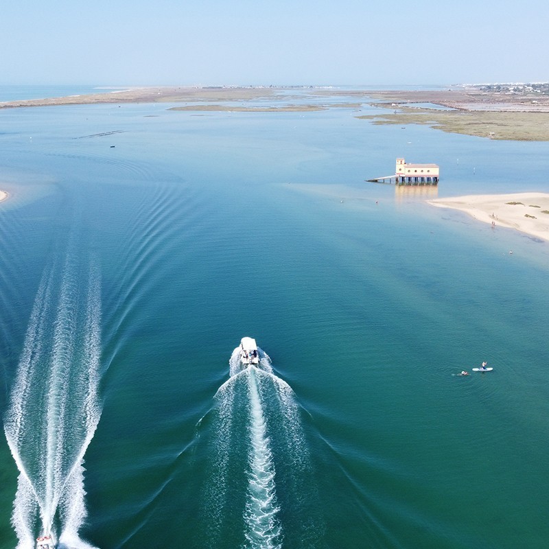 Vista aérea de um canal da Ria Formosa, com duas embarcações a deixar rasto de espuma e um edifício ocre sobre estacas na margem.