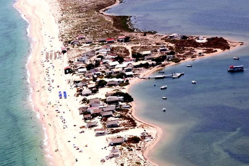 Vista aérea da ilha com dezenas de casas brancas aglomeradas entre a praia oceânica e a margem da Ria, com barcos fundeados.