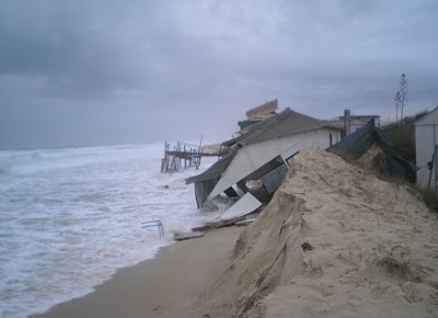 Mar revolto a galgar a duna sob céu cinzento, com casas de praia derrubadas e destroços de madeira espalhados na areia.
