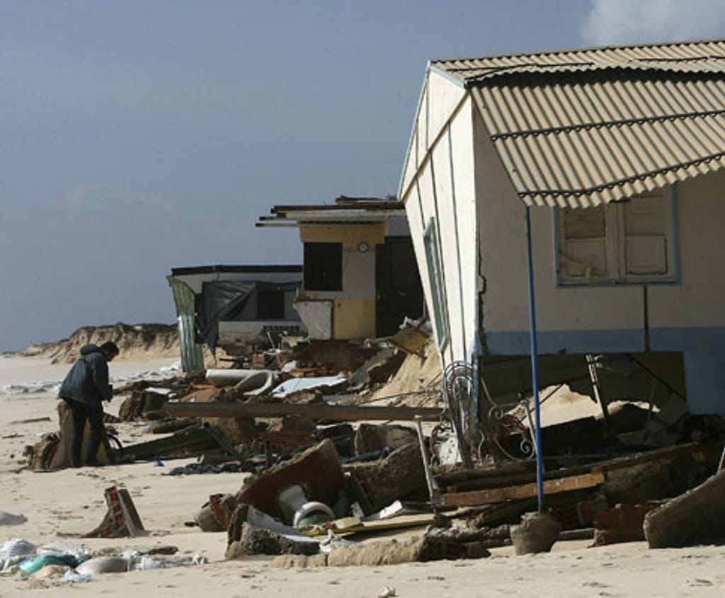 Homem entre destroços junto a casas de praia com as fundações escavadas pelo mar, portadas abertas e tijolos espalhados na areia.