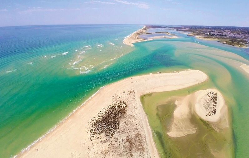 Vista aérea da Barra Nova: abertura no cordão de areia por onde o mar turquesa entra na Ria Formosa, entre línguas de areal.