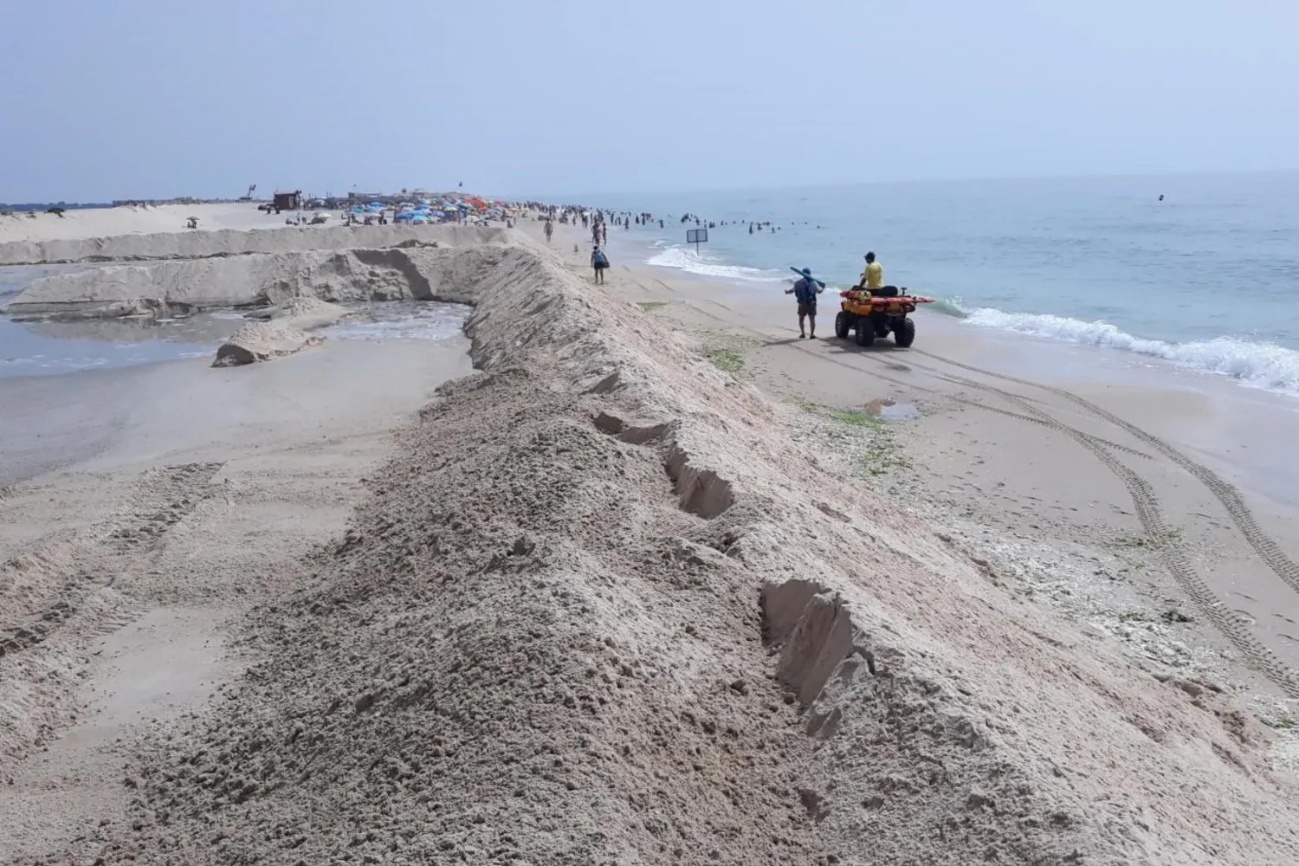 Monte alongado de areia recém-depositada ao longo da praia, com rastos de máquinas, um moto-quatro junto à água e banhistas ao fundo.