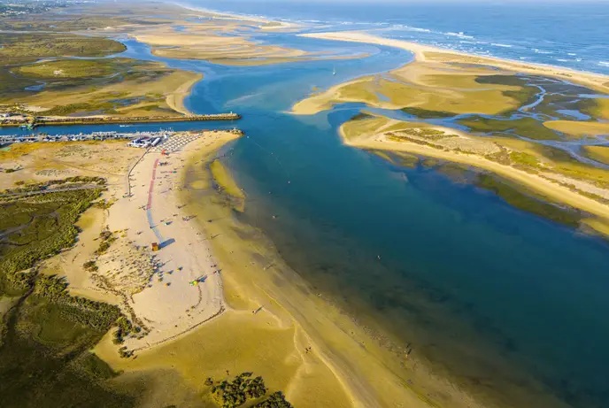 Vista aérea da Praia da Fuseta-Ria: língua de areia com chapéus de sol, cais com embarcações, canal turquesa da ria e ilha-barreira ao fundo