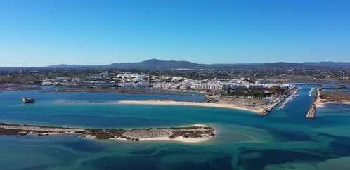 Vista aérea da Fuseta a partir da ria: casario branco, cais com embarcações, bancos de areia e canais turquesa, serra ao fundo