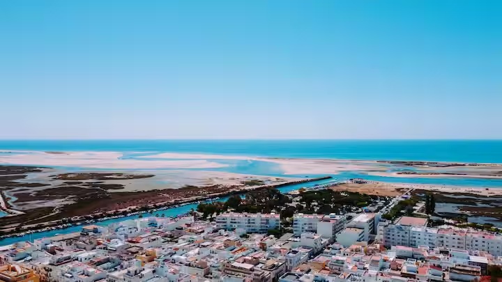 Vista aérea da Fuseta: casario branco em primeiro plano, canal com embarcações, ria com bancos de areia, ilha-barreira e oceano no horizonte