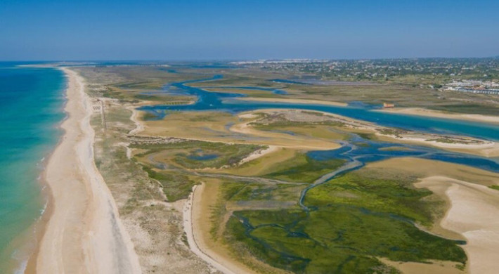 Vista aérea da Ria Formosa: praia e dunas da ilha-barreira à esquerda, canais serpenteantes entre sapais e bancos de areia, povoação ao fundo.