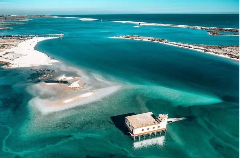 Vista aérea do Salva-Vidas da Fuseta sobre estacas na água turquesa da Ria Formosa, entre bancos de areia e ilhas-barreira.