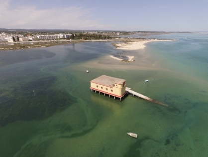 Vista aérea do Salva-Vidas sobre pilares na ria, com rampa, barcos pequenos e as casas brancas da Fuseta ao fundo.